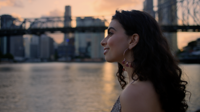 Brisbane Colourist Angela Cerasi did a warm grade on this shot of a smiling woman with dark curly brown hair looking out over the sunset-lit bay, a bridge and cityscape in the background, for the "Nothing's Changed. Everything's Different" campaign commercial for Brisbane Marketing